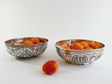 two silver bowls with oranges in them on a white surface