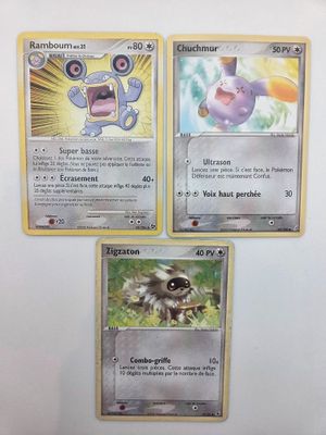 Trois cartes Pokémon sont posées sur une surface blanche bordée d'un liseré jaune.