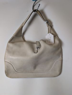 sac à main en cuir beige accroché à un mur blanc, avec une étiquette attachée dessus