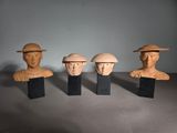 trois têtes d'hommes en bois coiffées de chapeaux, posées sur des socles sur une table