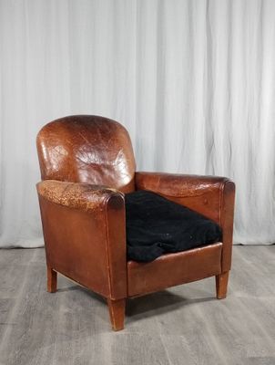 fauteuil en cuir marron avec un coussin noir, posé sur un parquet, devant un rideau blanc