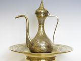 une théière en laiton ancien posée sur une assiette dorée à fond blanc