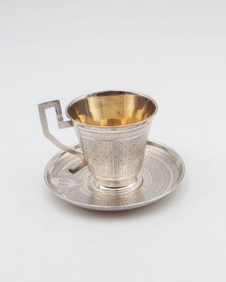 une tasse et une soucoupe anciennes en argent, posées sur une soucoupe, sur une surface blanche