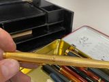 a person holding a gold colored pen in front of a box of pens