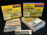 trois trains miniatures dans leurs boîtes, posés sur une surface noire, avec un pont en arrière-plan
