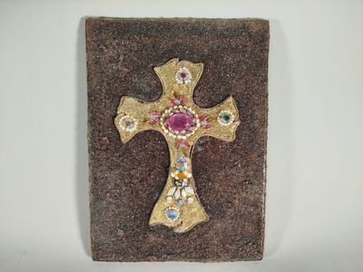 croix en or ornée de pierres roses et bleues, posée sur un socle en pierre marron