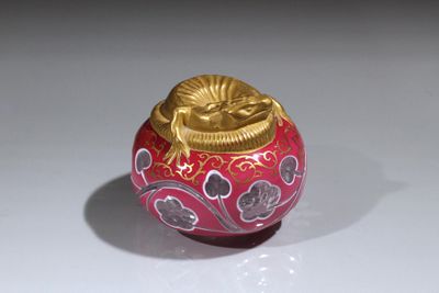 un vase rouge avec un couvercle doré posé sur une surface blanche