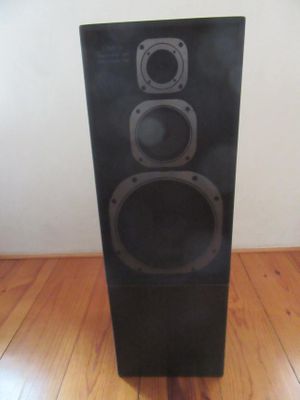 Une enceinte noire est posée sur un parquet, devant un mur blanc.