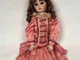 une poupée ancienne assise sur une surface blanche, vêtue d'une robe rose et de chaussures noires