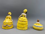 trois figurines en céramique représentant des femmes vêtues de robes jaunes et coiffées de chapeaux