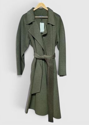 manteau en laine verte avec une ceinture, suspendu à un cintre contre un mur blanc