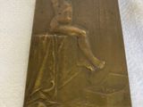 plaque en laiton représentant une femme assise sur une table, les jambes croisées