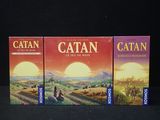 trois boîtes du jeu Catan posées sur une surface noire