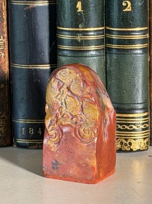 a red stone with a gold design on it in front of a row of books