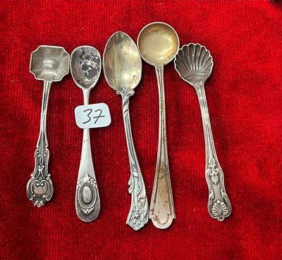 cinq cuillères en argent et une cuillère portant une étiquette avec un nom, posées sur un tissu rouge