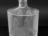 black and white photograph of a clear glass bottle with three figures on it