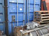 a large stack of metal barrels sitting on a pallet in front of a blue shipping container