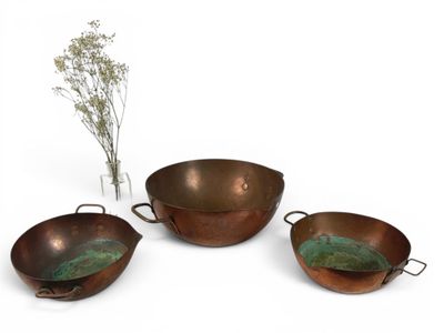 trois casseroles en cuivre et un vase avec des fleurs, posés sur une surface blanche