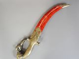 une image représentant un couteau en forme de lézard, de couleur rouge et or, posé sur une surface blanche