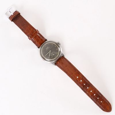La montre repose sur une surface blanche et est dotée d'un bracelet marron.