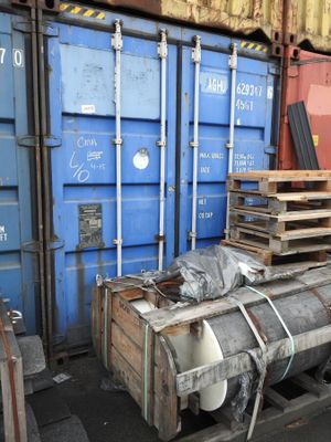 a large stack of metal barrels sitting on a pallet in front of a blue shipping container