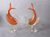 une paire de sculptures de poissons en verre orange posées sur des socles transparents, sur fond blanc