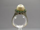 une image représentant une bague ornée d'une perle blanche et d'une émeraude, posée sur une surface transparente