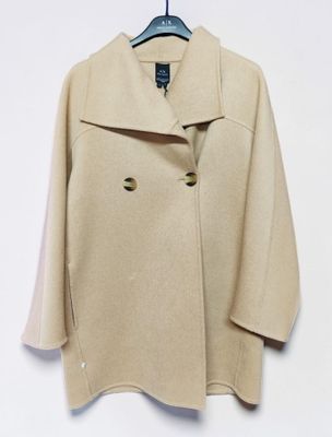manteau en laine beige suspendu à un cintre devant un mur blanc, sur fond blanc
