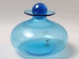 un vase en verre bleu avec un couvercle bleu posé sur une surface blanche
