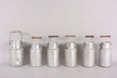 six metal milk cans with wooden handles lined up against a white wall
