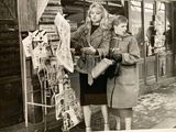 two women standing outside of a store looking at newspapers
