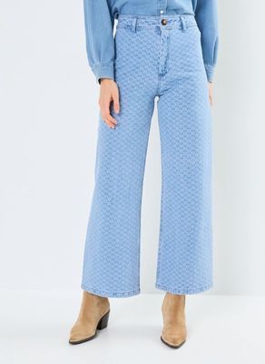 Une femme vêtue d'une chemise bleue et d'un jean se tient debout devant un fond blanc.