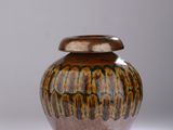 un vase marron et noir muni d'un couvercle, posé sur une surface blanche