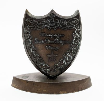 un bouclier antique en bronze posé sur un socle en bois, sur fond blanc