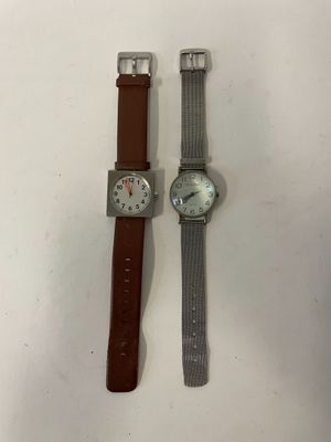 deux montres posées sur une surface blanche : l'une est dotée d'un bracelet marron et l'autre est une montre argentée avec un cadran argenté