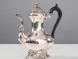 une théière ancienne en argent, avec une anse noire, posée sur une table blanche