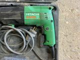 a green drill in a case on the floor next to a pair of brown shoes