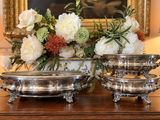 deux plats en argent posés sur une table, avec des fleurs en arrière-plan