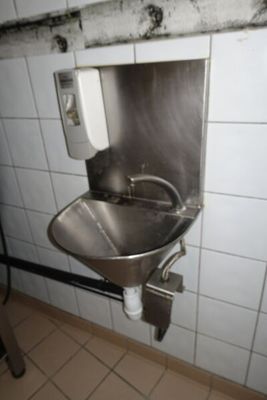 un lavabo en acier inoxydable dans des toilettes publiques, avec un distributeur de savon fixé au mur