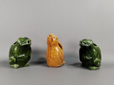 trois figurines en céramique représentant des grenouilles et un oiseau, posées sur un fond gris