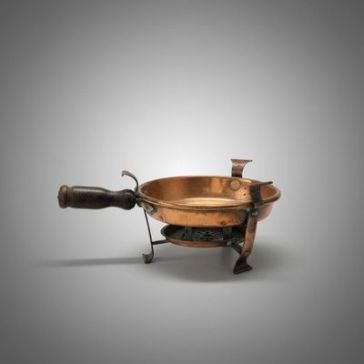 une vieille casserole en cuivre avec une poignée en bois, posée sur un support