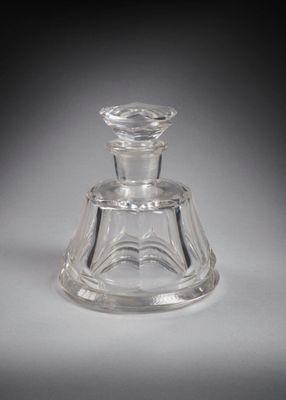 un flacon de parfum en verre à l'ancienne, avec un socle transparent posé sur une surface blanche