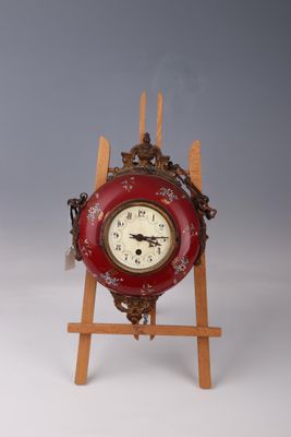 une petite horloge rouge posée sur un chevalet en bois, sur un fond gris