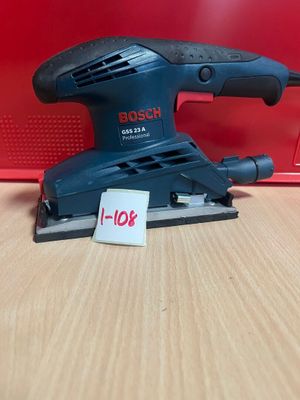 une ponceuse orbitale Bosch posée sur une table en bois, à côté d'une boîte rouge