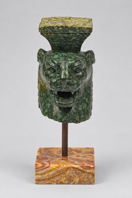 une photo représentant une sculpture verte représentant une tête, posée sur un socle en bois