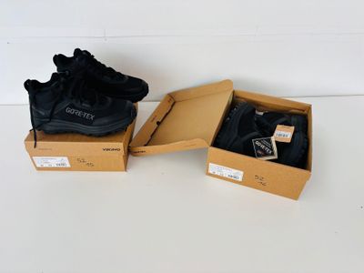 Deux paires de baskets noires se trouvent dans une boîte posée sur une surface blanche.