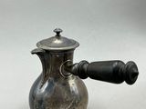une cafetière ancienne en argent, dotée d'une poignée en bois, posée sur un support blanc