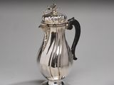 une cafetière ancienne en argent avec couvercle, posée sur un socle