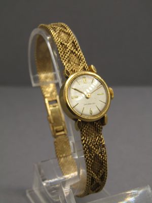 une photo d'une montre en or avec un bracelet maillé, posée sur un socle transparent