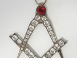 une image représentant un pendentif maçonnique en forme d'équerre et de compas, orné d'une pierre rouge